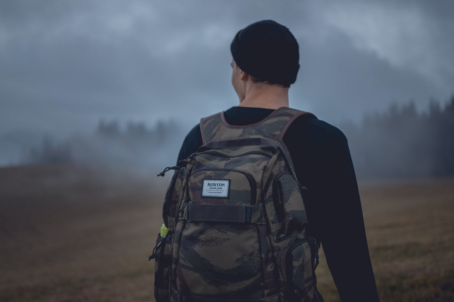 man in black shirt and black knit cap, wearing brown and green camouflage backpack standing on brown field